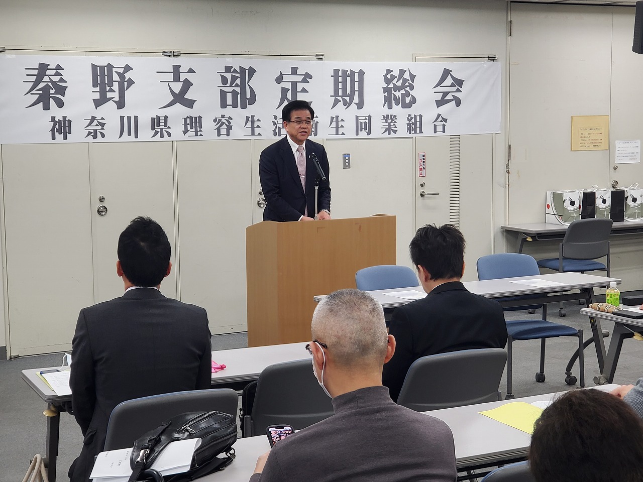 「秦野支部定期総会」と書かれた横断幕が掲げられた会場で、スーツ姿の市長が演台から挨拶し、参加者が聞いている様子の写真