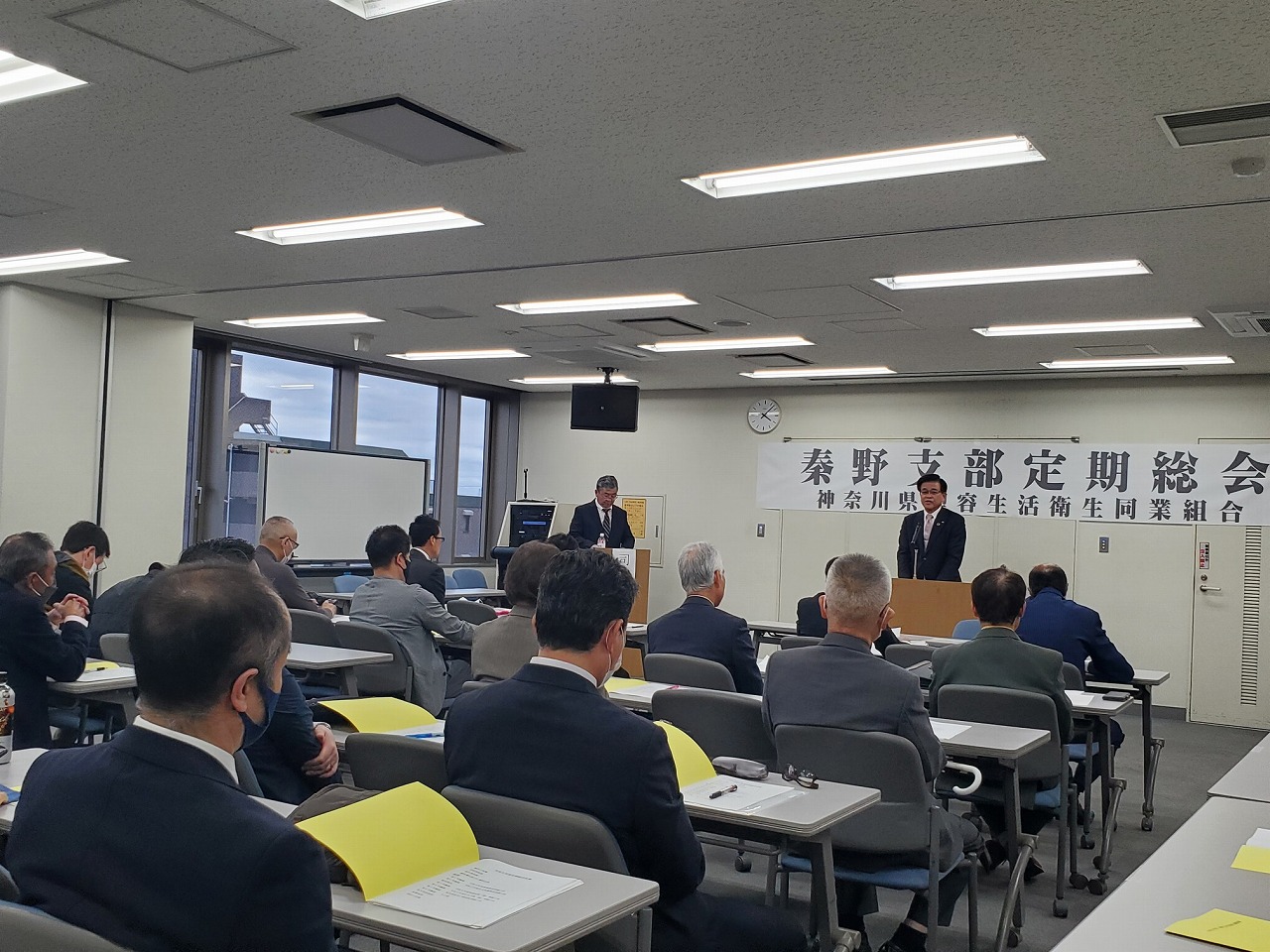 「秦野支部定期総会」の横断幕が掲げられた会場で、演台に立つ市長が挨拶し、演台の前方に設置されたテーブル席に大勢の参加者が座り聞いている様子を後方から写した写真