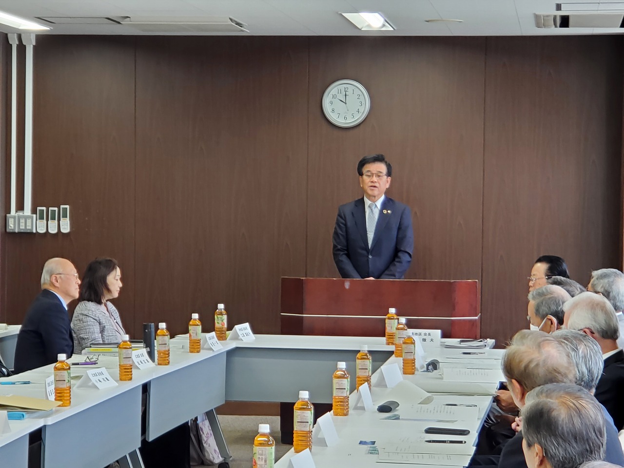 スーツ姿の市長が演台に立って挨拶し、設置された机に座り参加者たちが聞いている様子の写真