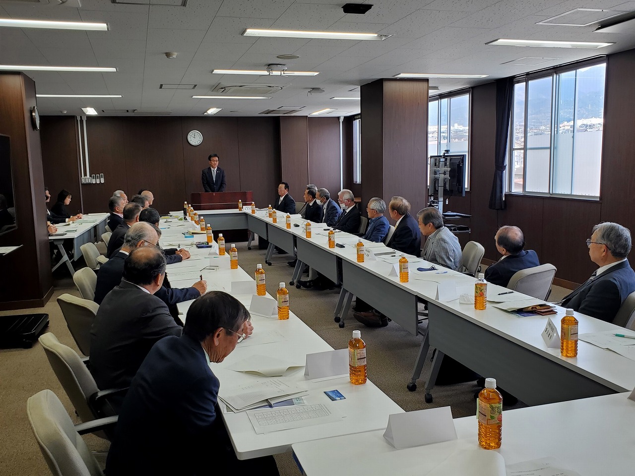 ロの字型にテーブルが置かれた広い会議室で、スーツ姿の市長が演台から話し、大勢の参加者が熱心に耳を傾けている様子を後方から写した写真