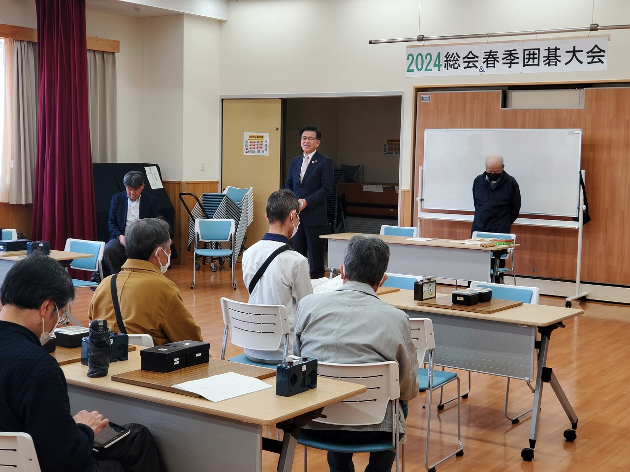 「春季囲碁大会」と書かれた横断幕が掲げられた会場の前方でスーツ姿の市長が挨拶している様子を、前方に設置された机に座り参加者の方々が聞いている様子の写真