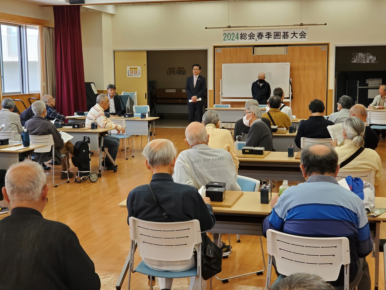 「春季囲碁大会」と書かれた横断幕が掲げられた会場の前方でスーツ姿の市長が挨拶している様子を、前方に設置された机に座り参加者の方々が聞いている様子の室内全体を写した写真