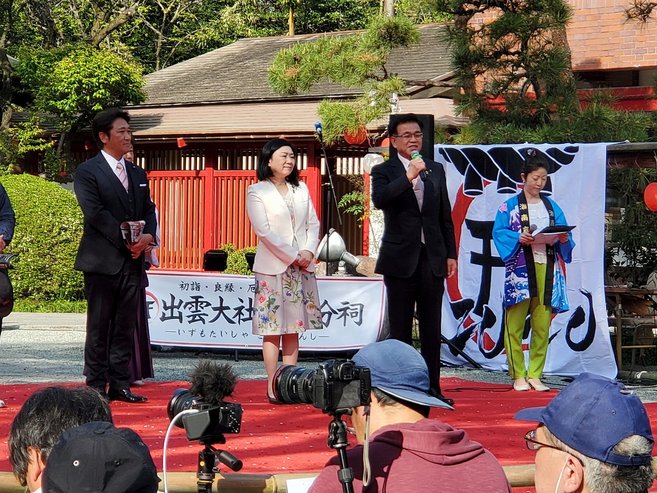 「出雲大社」と書かれた幕の前で、スーツ姿の市長がマイクで挨拶し、その様子を壇上の関係者や客席の観客が聞いている様子の写真