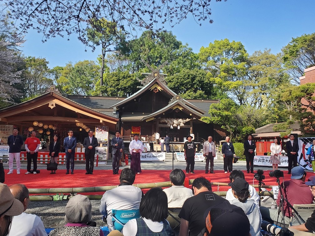 神社の社殿を背景に、赤い絨毯が敷かれたステージに来賓の方々が横一列に並び、それを見るために集まった観客が座っているを後方から写した写真