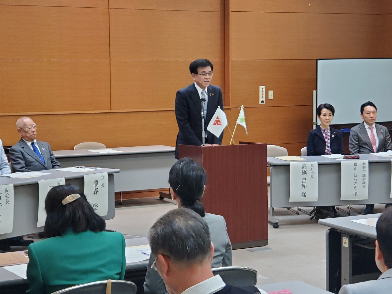 総会が開かれている会場で、スーツの市長が演台に立ちスピーチし、それを来賓や参加者が聞いている様子の写真