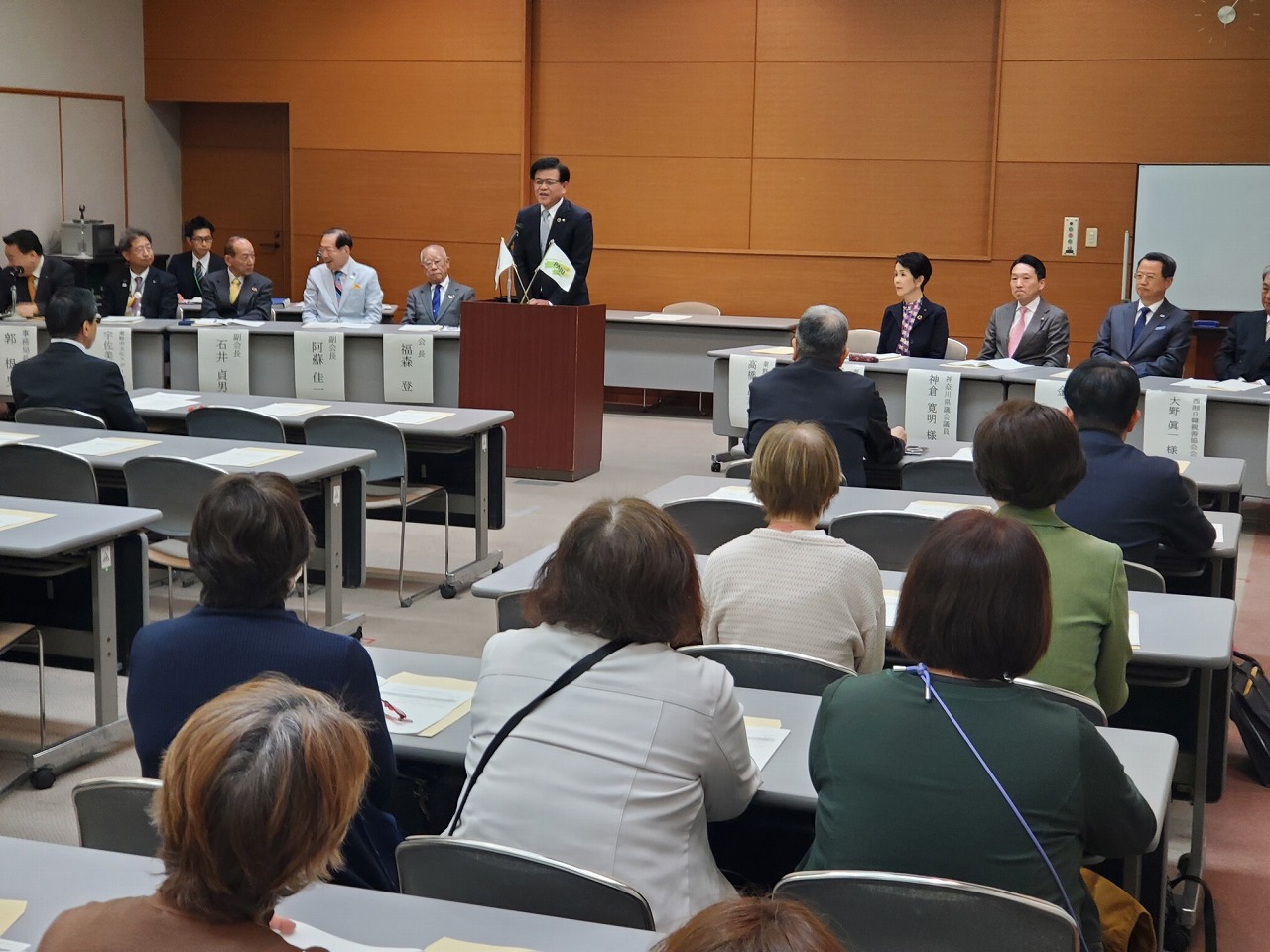 会場の前方にある演台に立ち市長がスピーチし、前方の来賓席と、後方のテーブル席に座った参加者が聞いている様子を後方から写した写真