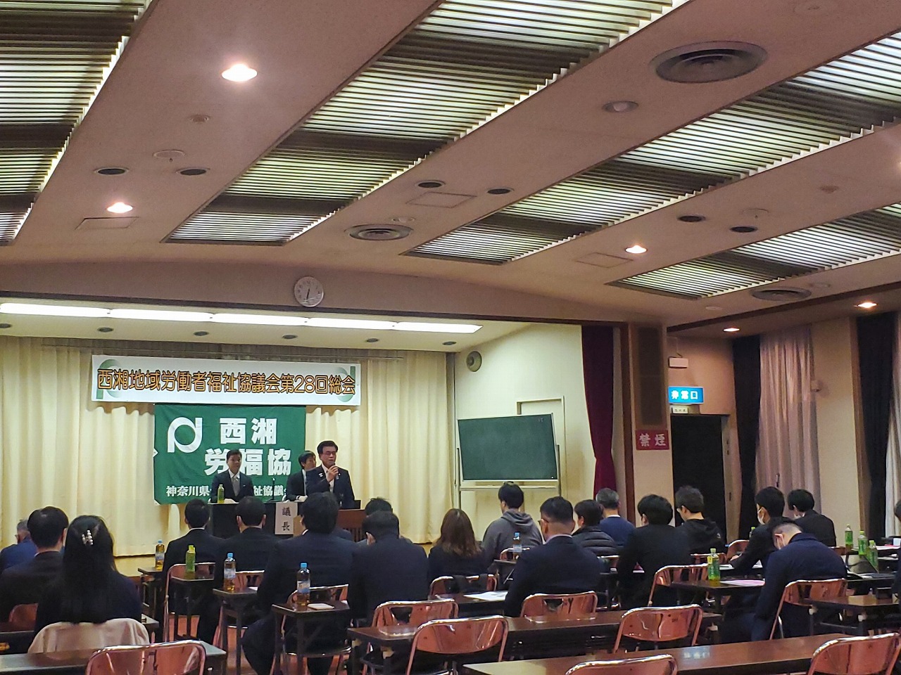 「第28回総会」の横断幕が掲げられた広い会場で、前方の壇上で市長が挨拶し、客席の多くの参加者がそれを聞いている様子を後方から写した写真