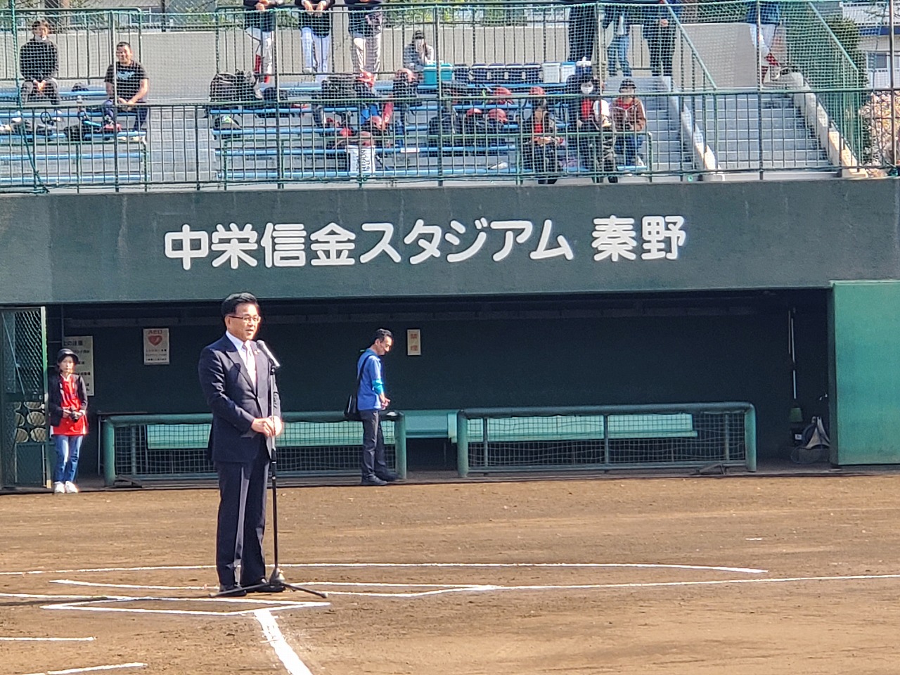「中栄信金スタジアム 秦野」野球場で、ホームベースのところに設置されたスタンドマイクの前に立ったスーツ姿の市長がマイクの前で挨拶している写真。