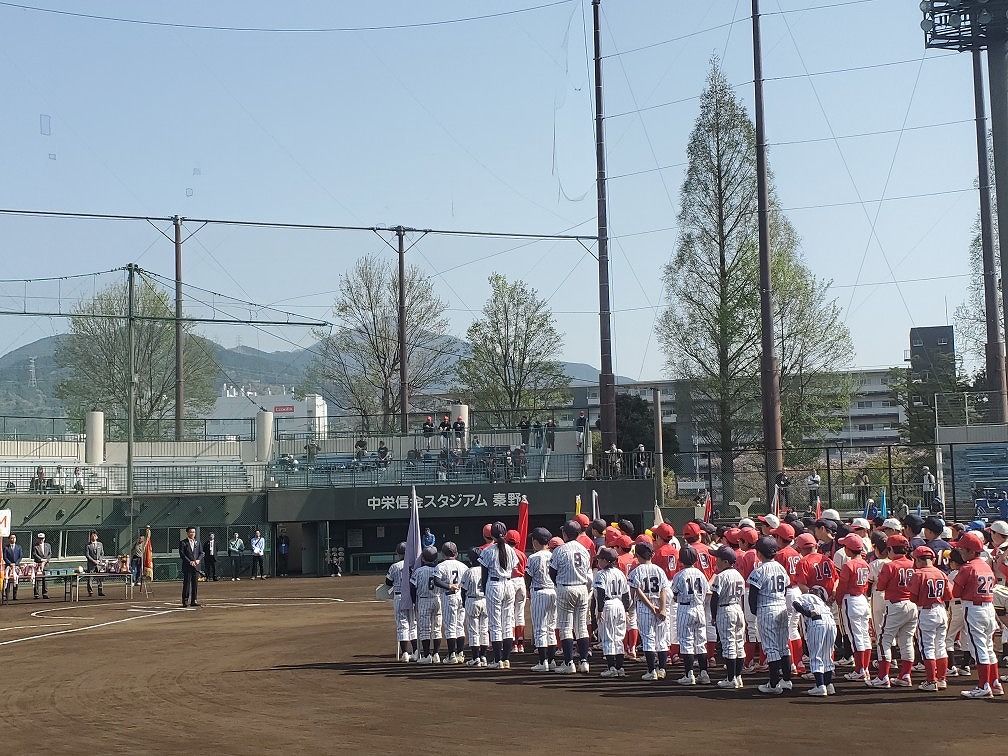 「中栄信金スタジアム 秦野」のグラウンドで、2つの少年野球チームのユニフォーム姿の選手たちが整列し、大会の開会式に臨んでいる写真