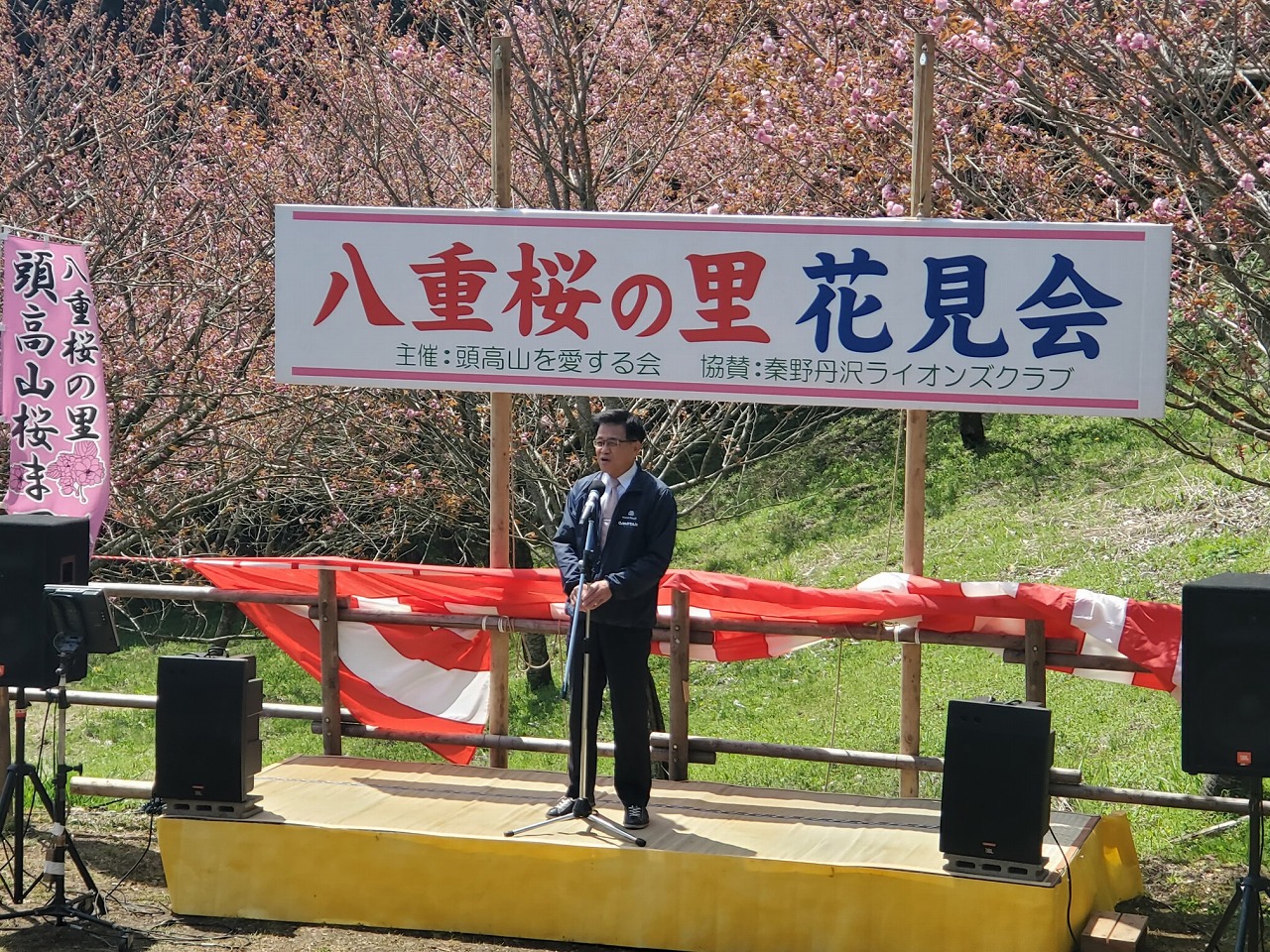 「八重桜の里 花見会」と書かれた横断幕が掲げられた屋外ステージで、満開の桜を背景に、市長がマイクの前で挨拶している写真