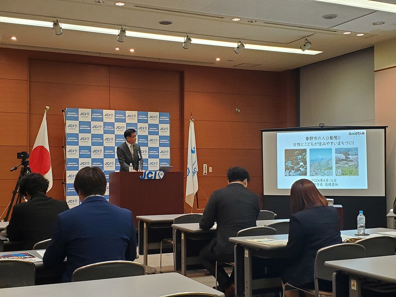 JCIのロゴがある背景の前に設置された演台にてスーツの市長が横のスクリーンに映しだされた資料を見ながら話している様子の写真