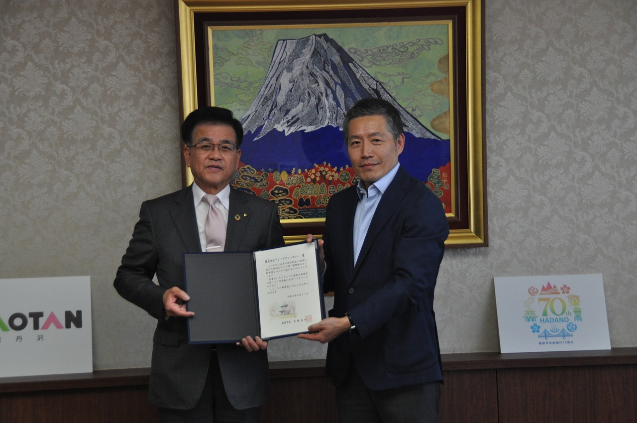 スーツ姿の市長と安川代表取締役社長が、二人で賞状のようなものを持ち、カメラに向かって微笑んでいる写真