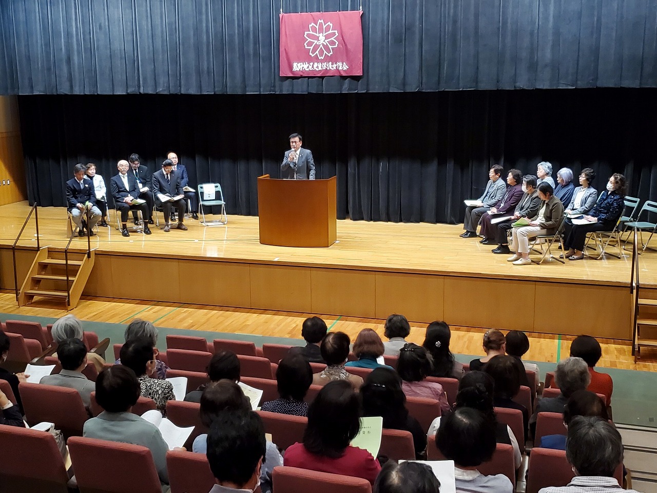 「更生保護女性会」の赤い旗が掲げられた講堂のステージで、演台の前で市長が挨拶し、壇上の来賓と客席に座る大勢の参加者が聞いている様子を後方から写した写真