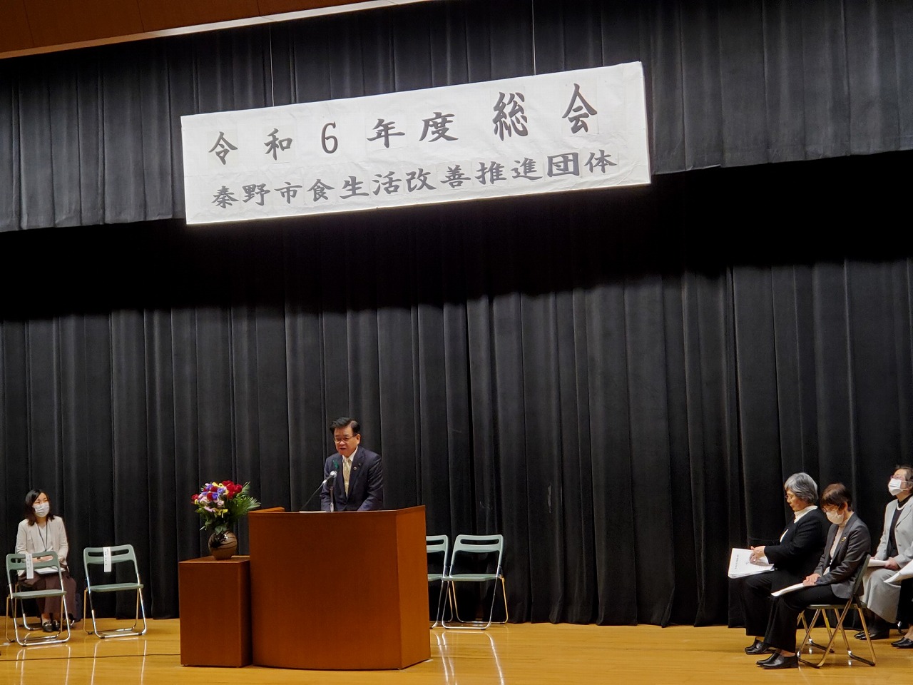 「令和6年度 総会」と書かれた横断幕のステージ上の演台でスーツ姿の市長が挨拶し、周りに座った関係者が話を聞いている写真