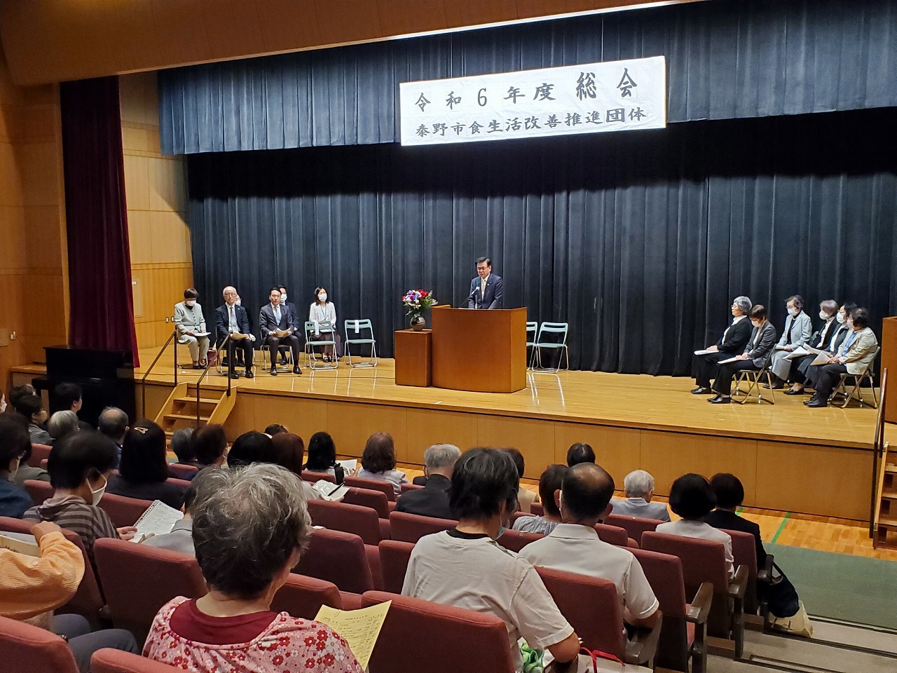 講堂のステージで「総会」が開かれ、演台に立つ市長の挨拶を、壇上の来賓と、客席を埋めた多くの聴衆が聞いている様子の写真