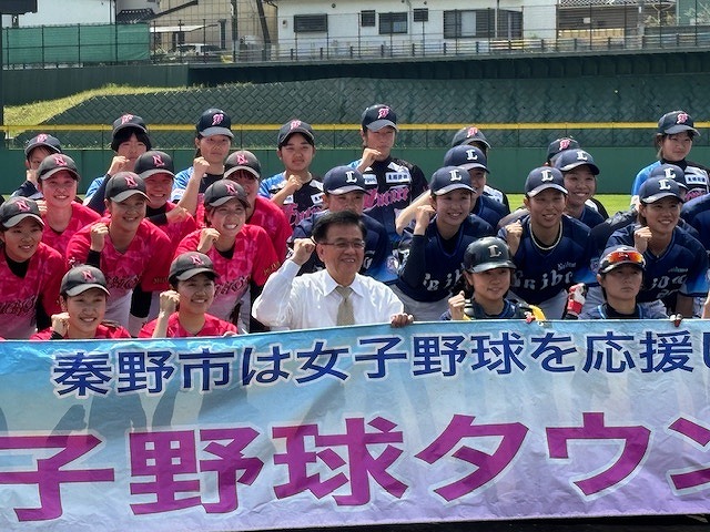 中央でガッツポーズをする市長をユニフォーム姿の大勢の女子野球選手が囲み、「秦野市は女子野球を応援」と書かれた横断幕を掲げて集合写真を撮っている写真
