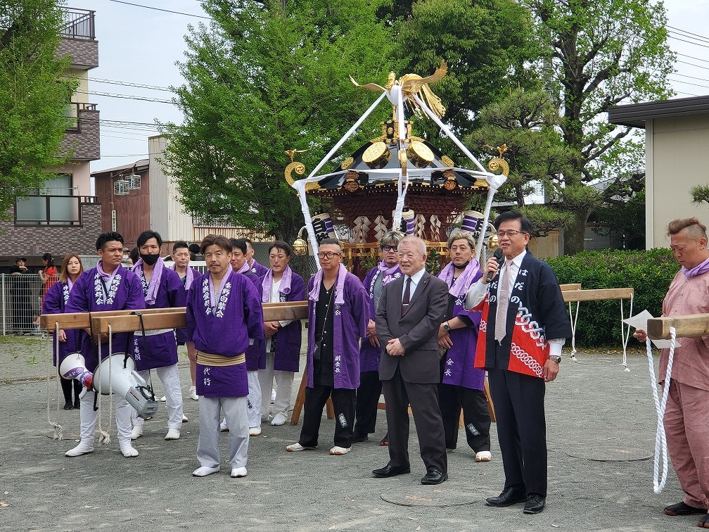 鳳凰が飾られた神輿を紫の法被を着た担ぎ手たちが囲み、スーツ姿の男性の横で法被姿の市長がマイクを持ち話している様子の写真