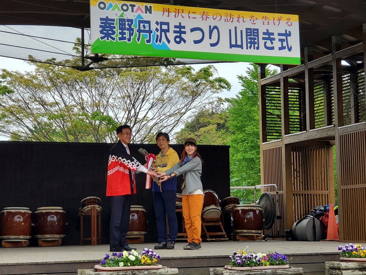 「秦野丹沢まつり 山開き式」の横断幕が掲げられた屋外ステージで、法被姿の市長が男女のペアに記念品のようなものを贈呈している写真