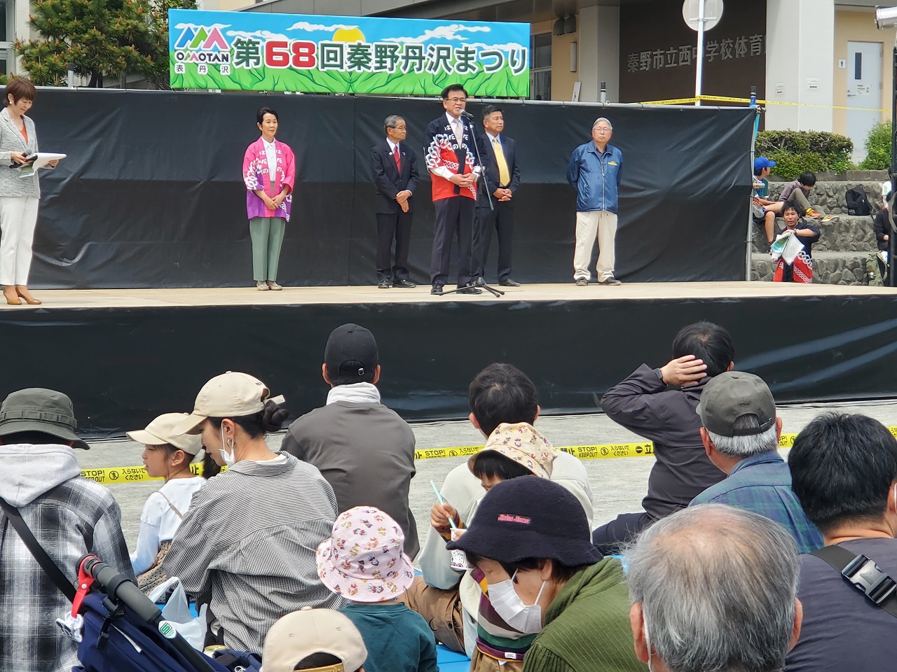 「第68回秦野丹沢まつり」と書かれた横断幕が設置されたステージ上で法被を着た市長が前方に座る大勢の参加者に向かい挨拶している様子を後方から写した写真