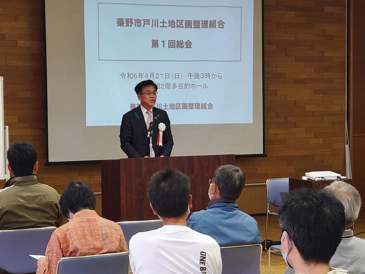 資料が映し出されたスクリーンを背に、スーツ姿の市長が演台でスピーチを行い、椅子に座った数名の参加者がそれを聞いている様子の写真