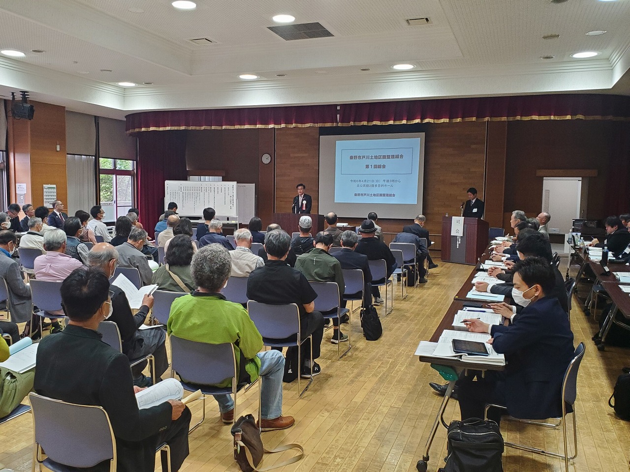 資料が映し出された広い会場で、壇上に立つ市長が挨拶し、それを椅子に並んで座る大勢の参加者が視聴している様子を後方から写した写真