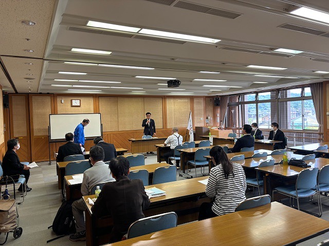 研修会のような会場で、前方に立つスーツ姿の市長が話す横で、青い上着を着た男性がお辞儀をするのを、机に向かって着席している十数名の参加者が見ている様子の写真