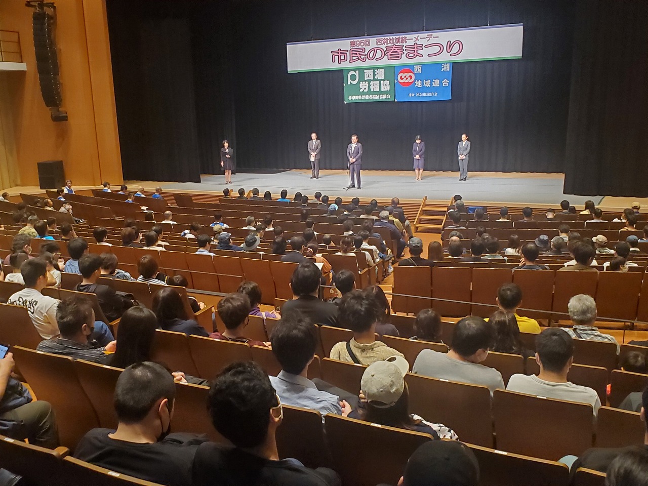 「市民の若者まつり」と書かれた横断幕が掲げられたステージ上で数名の登壇者が並び、中央のマイクの前に立ち市長が挨拶をする様子を、客席にいる大勢の観客が聞いている様子を後方から写した写真