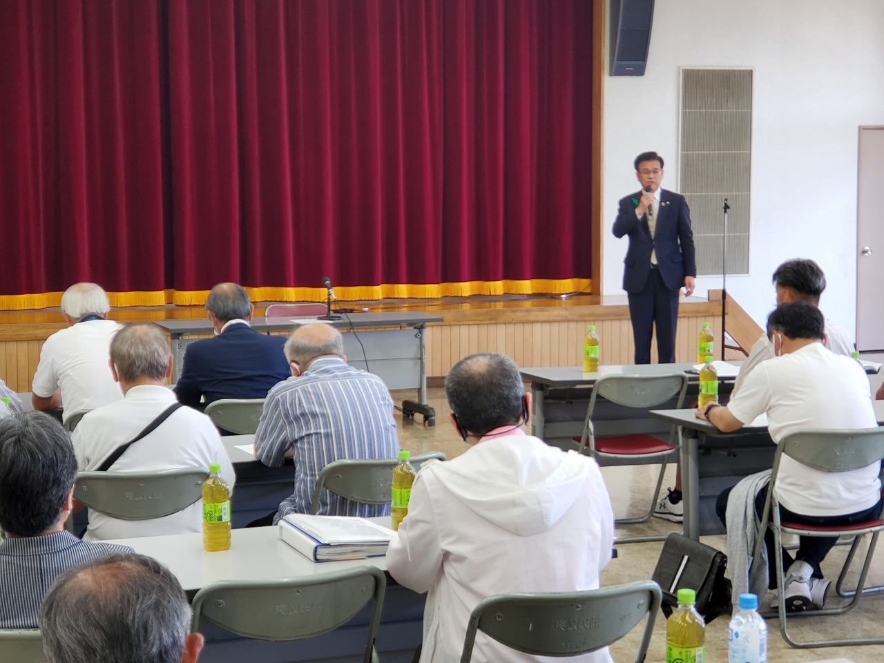 赤い緞帳のが下りている舞台の前でスーツ姿の市長がマイクを持って話しており、その前方に並べられた机に着席した多くの聴衆が耳を傾けている講演会の写真