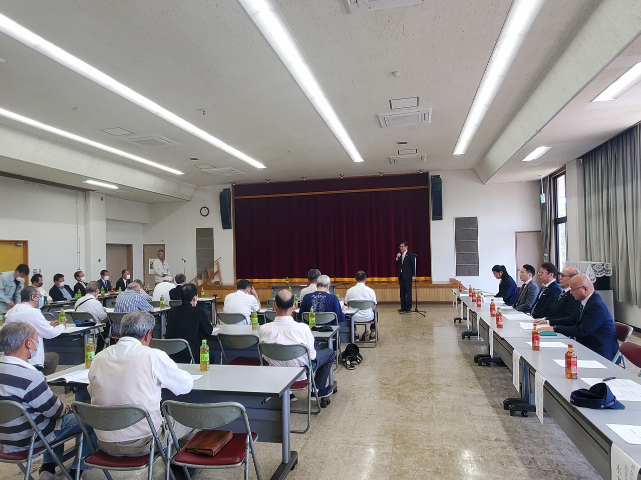 ステージ前でスーツ姿の市長が、会場内の長机に着席した多くの参加者に向けて話している様子を後方から写した写真