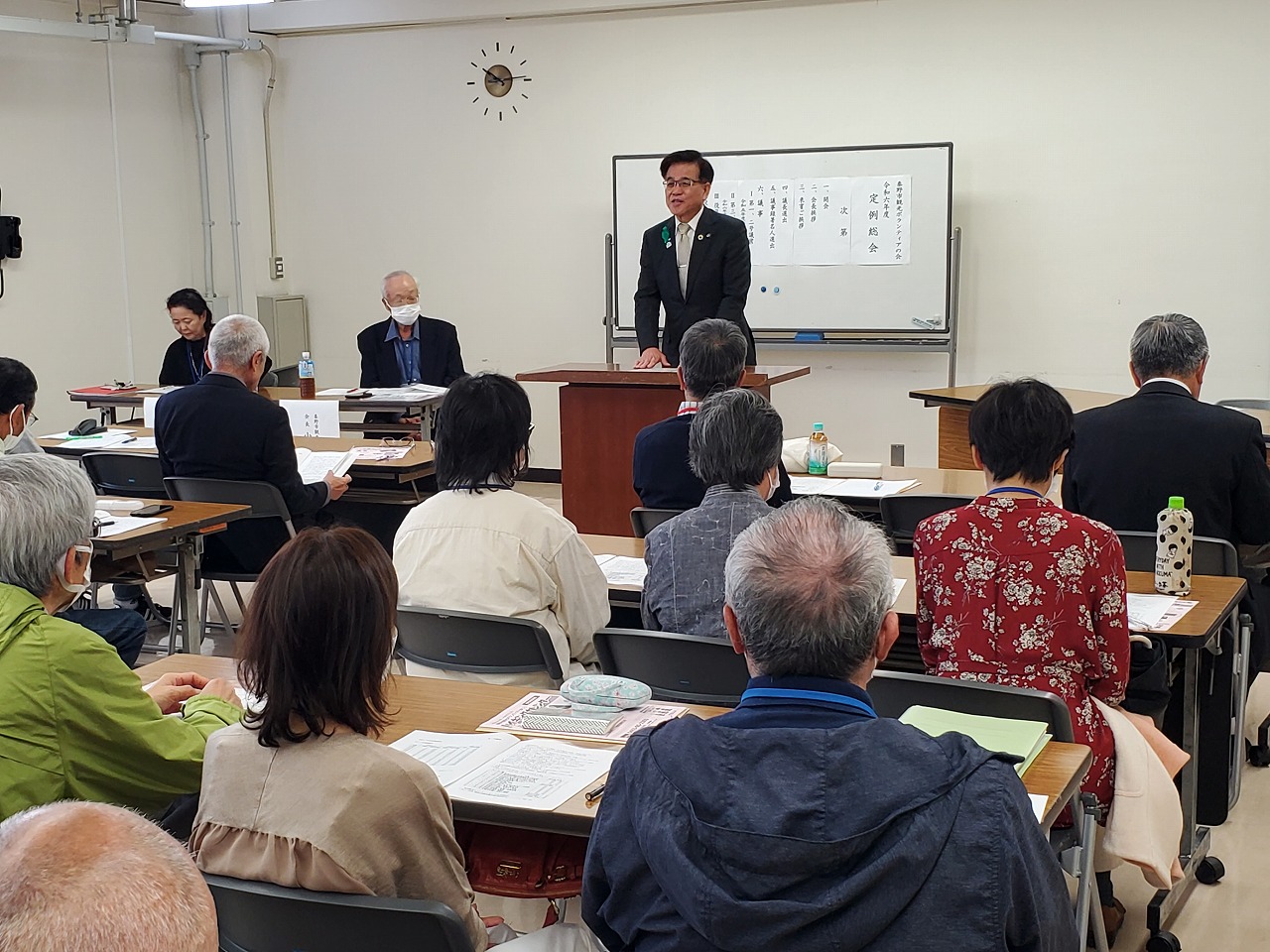 ホワイトボードの前に設置されてた演台の前にスーツ姿の市長が立ち、前方の参加者に向かって話をしている様子の写真