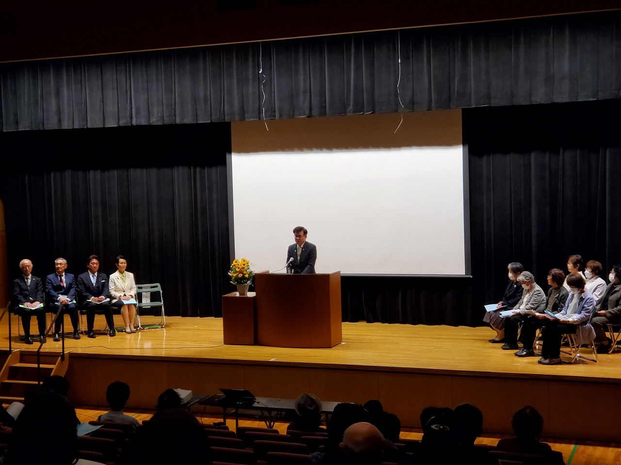 講堂のステージ上で市長が演台に向かってスピーチを行い、その両脇には複数の来賓が着席し、客席の聴衆が聞いている様子の写真