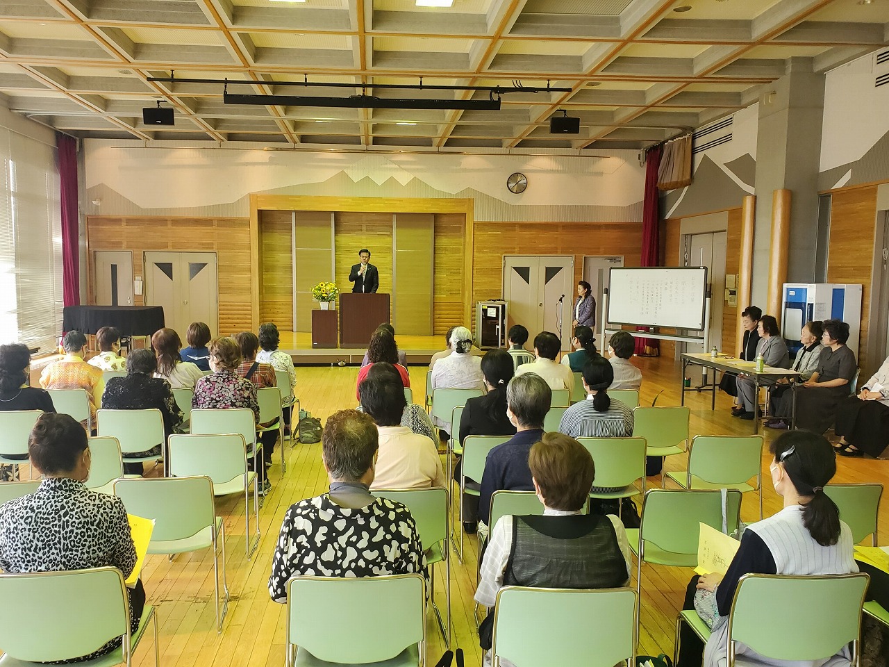 前方に設置された演台の前でスーツ姿の市長がマイクを持って挨拶し、整然と椅子に座る多くの参加者が真剣に耳を傾ける様子を後方から写した写真