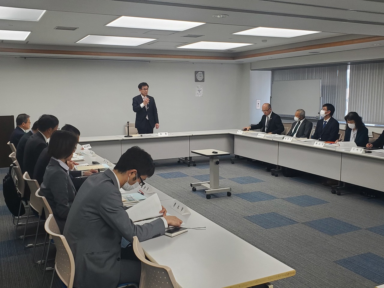 テーブルが置かれた会議室で、スーツ姿の市長がマイクを手に立って発言し、他の参加者たちが着席して資料に目を通したり話を聞いたりしている様子の写真