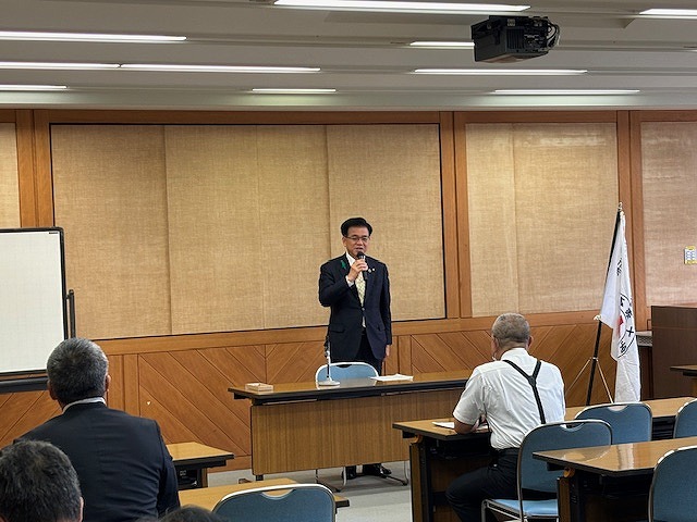 前方に設置された机の前にスーツ姿の市長がマイクを持ち立って話をして言う様子を、机に向かって座る参加者たちが耳を傾けている様子の写真