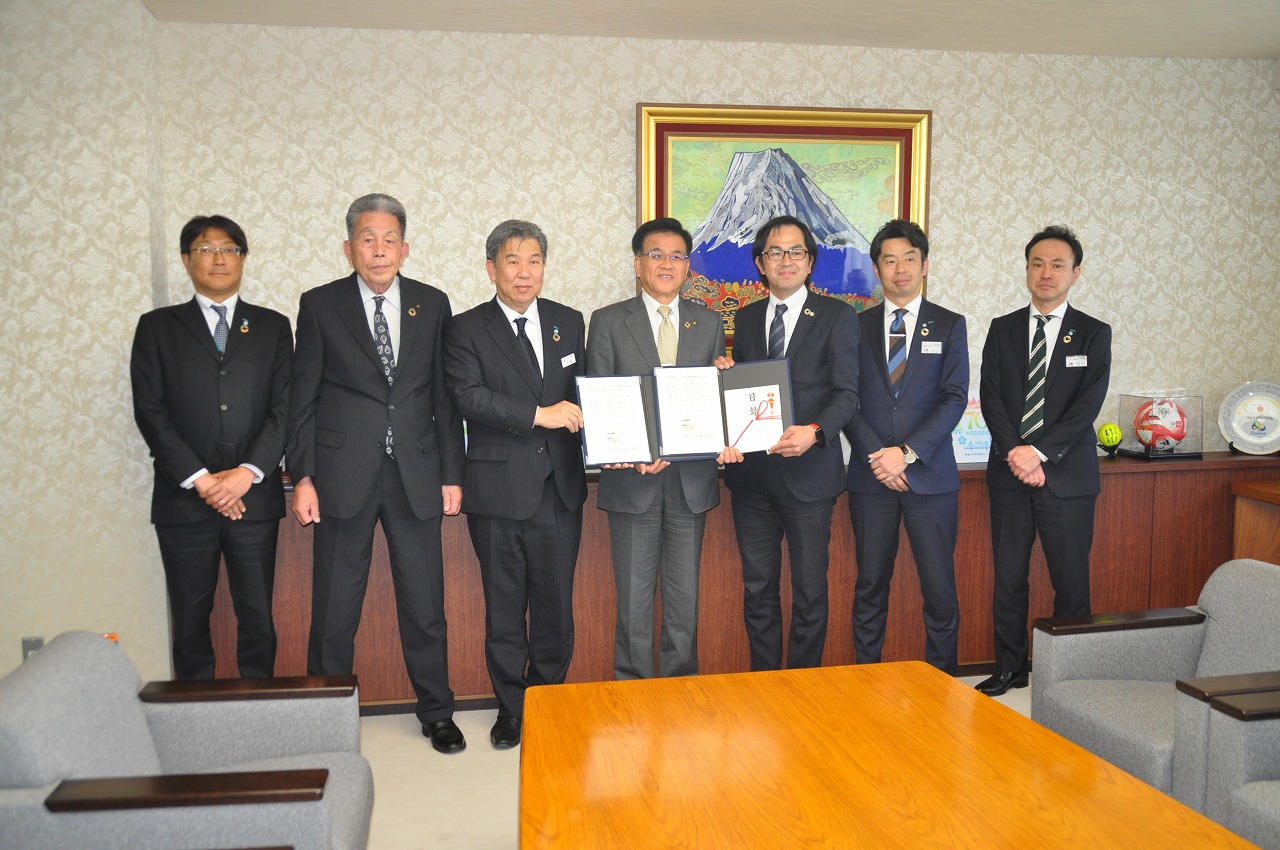 スーツ姿の男性7名が横一列に並び、中央に市長とその両サイドに男性と一緒に目録などを手に持って記念撮影をしている写真