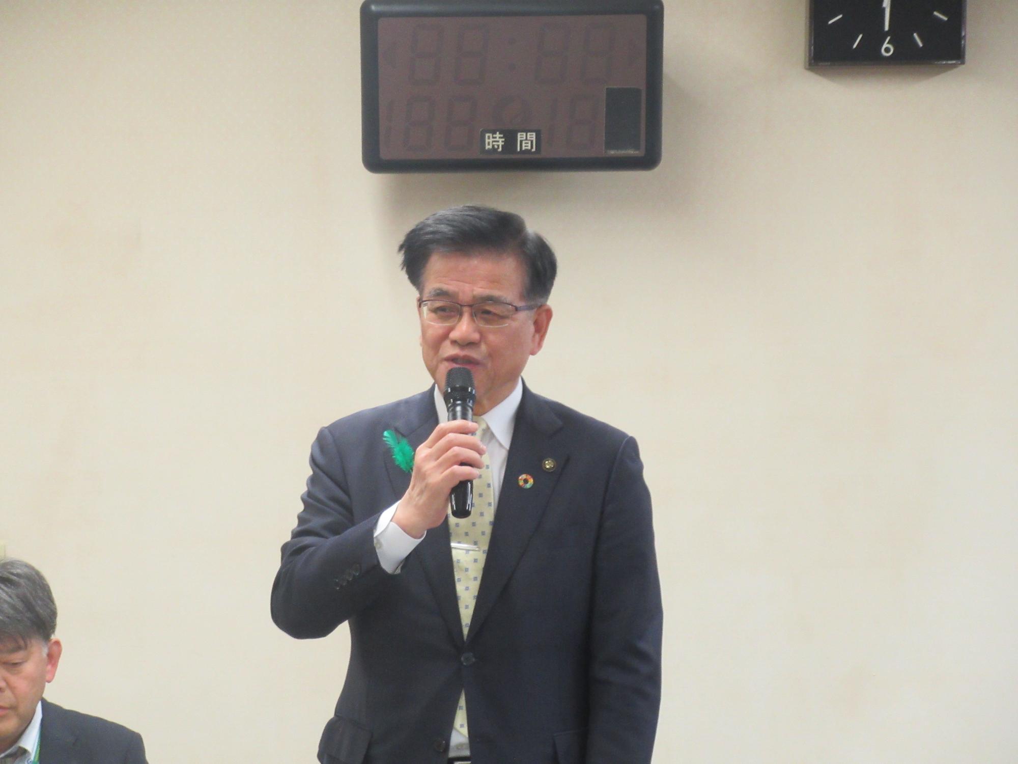 胸に緑色の羽根を付けたスーツ姿の市長が、マイクで話をしている様子の写真