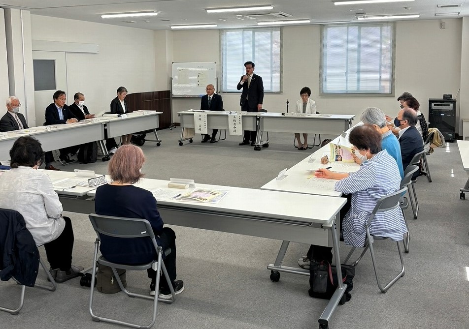 ロの字型にテーブルが配置された会議室で、スーツ姿の市長がマイクで話し、周りに座った多くの参加者が資料を見ながら聞いている様子を後方から写した写真