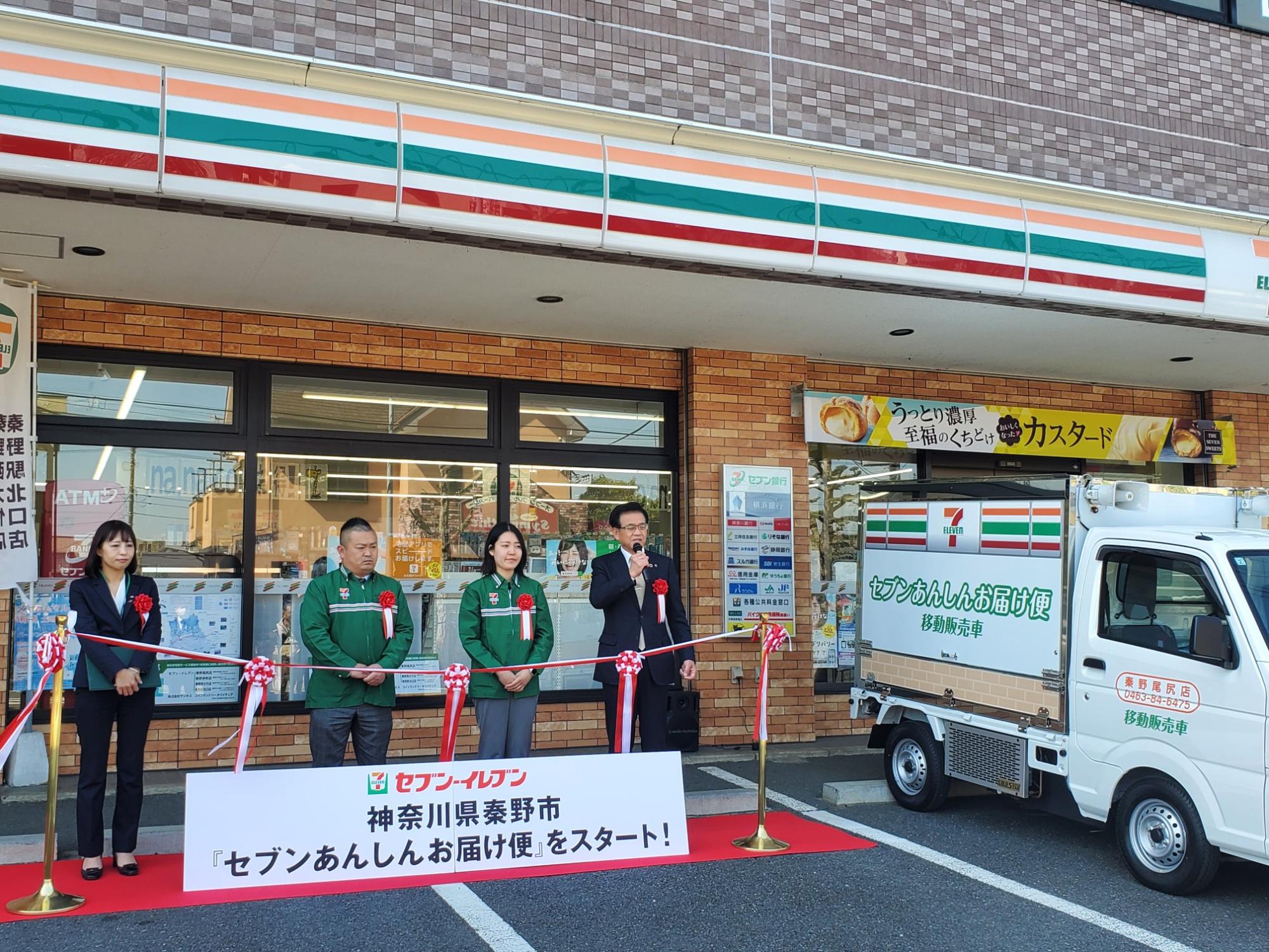 セブンイレブンの店先で「セブンあんしんお届け便」の開始セレモニーが開かれ、移動販売車の横でスーツの市長が挨拶している写真