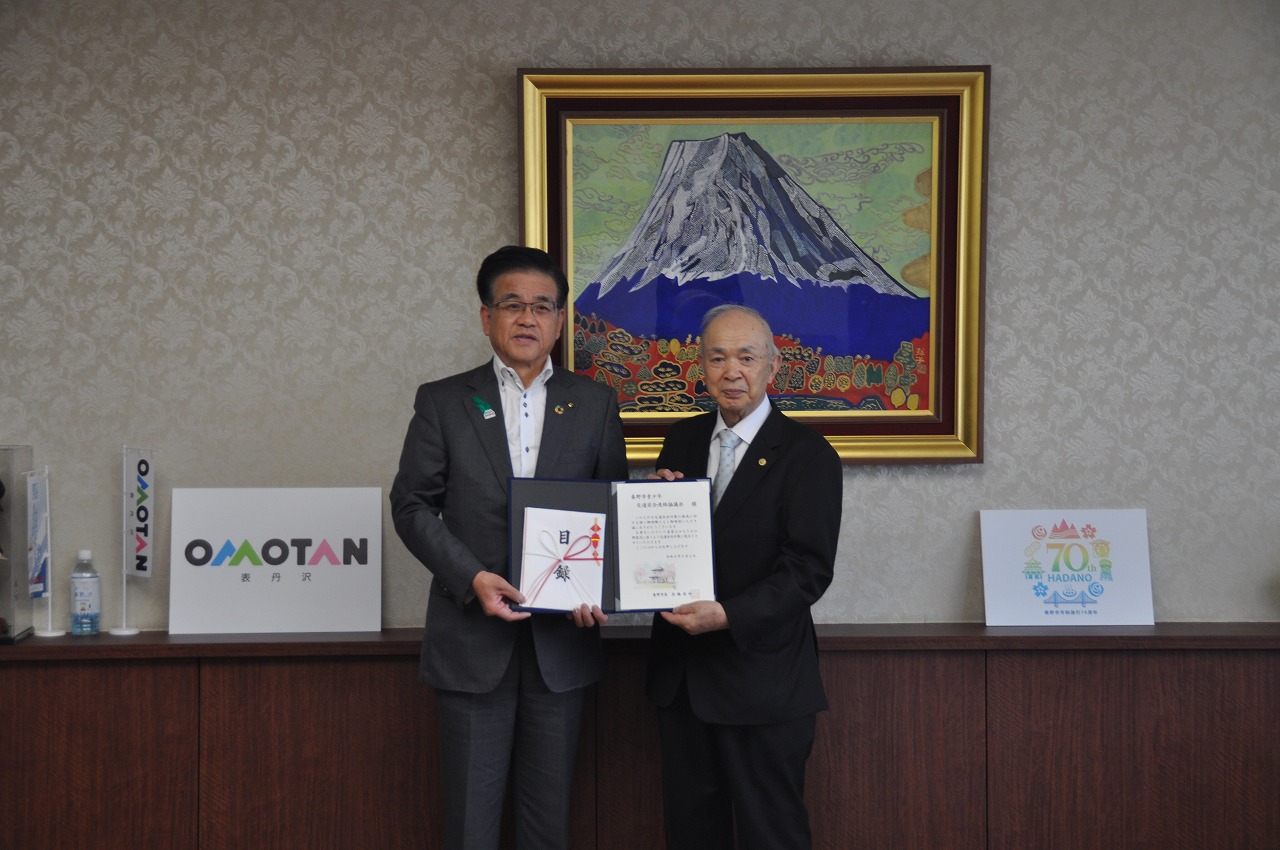 壁に飾られた富士山の絵を背景に、スーツ姿の市長と川口会長が賞状と目録を持って並んで立っている様子の写真