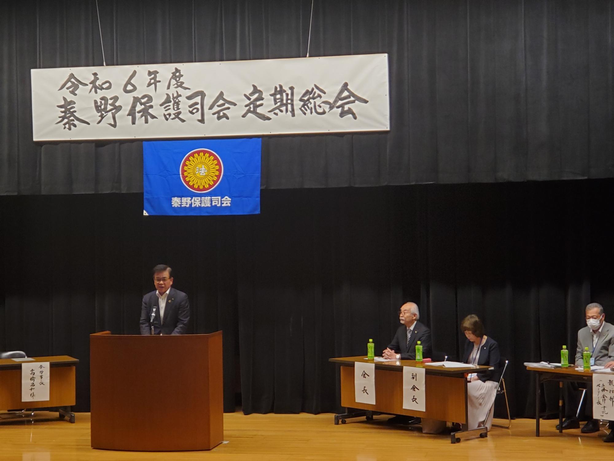 「令和6年度 秦野保護司会定期総会」と書かれた横断幕が掲げられたステージ上で、スーツを着た市長が演台からスピーチをしており、複数の人々が机に着席している様子の写真