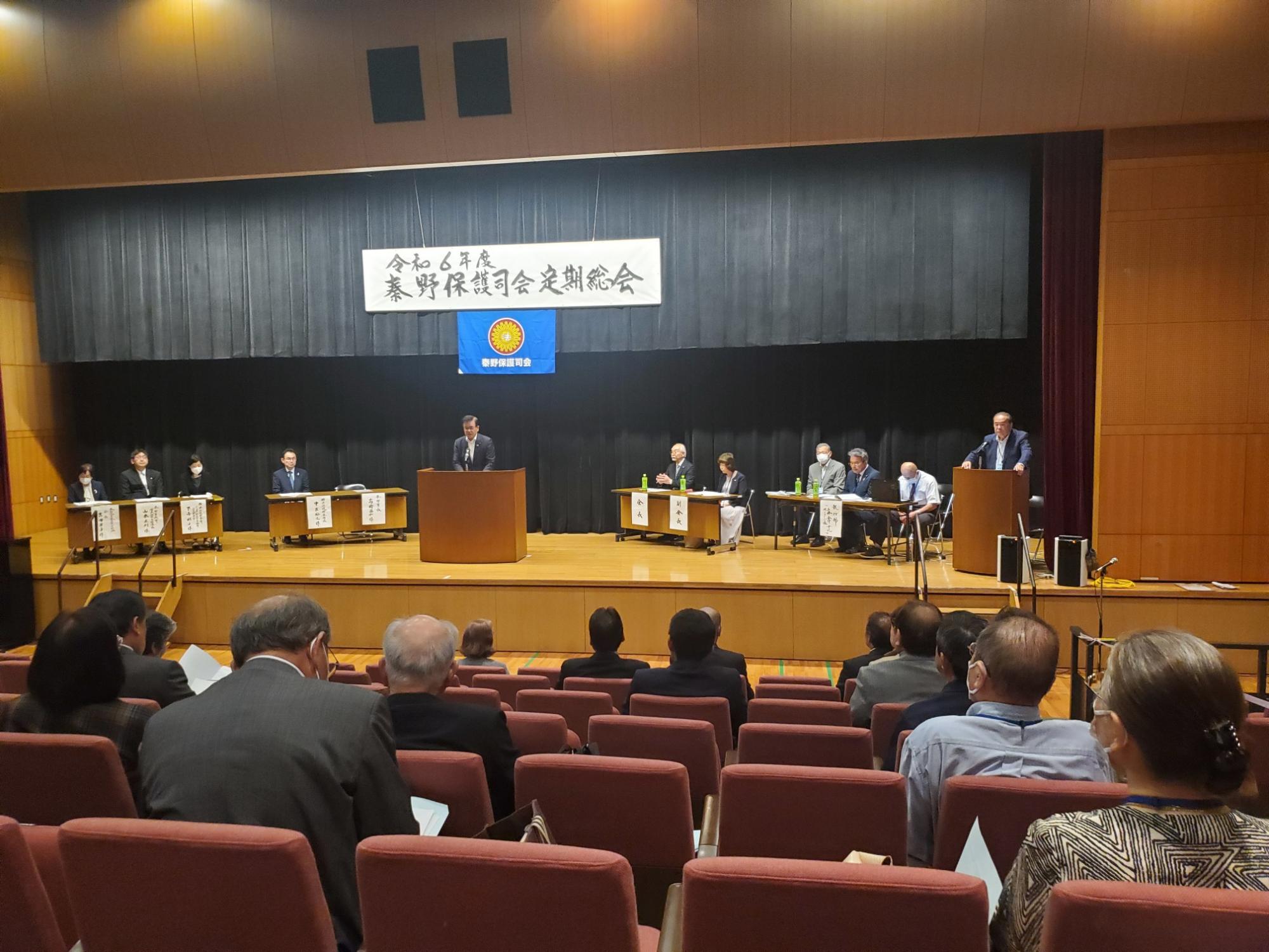 「令和6年度 秦野保護司会定期総会」と書かれた横断幕が掲げられたステージ上で、スーツを着た男性が演台から大勢の聴衆と壇上の人々にスピーチをしている様子の写真