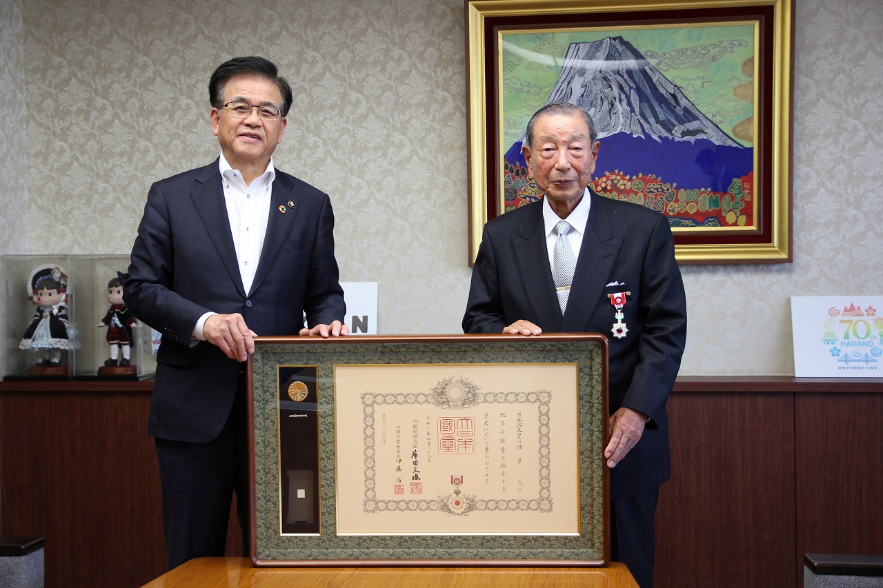 複数のテーブルと椅子が並んだ広間で、スーツ姿の市長が演台から大勢の聴衆に向かってスピーチをしている様子の写真