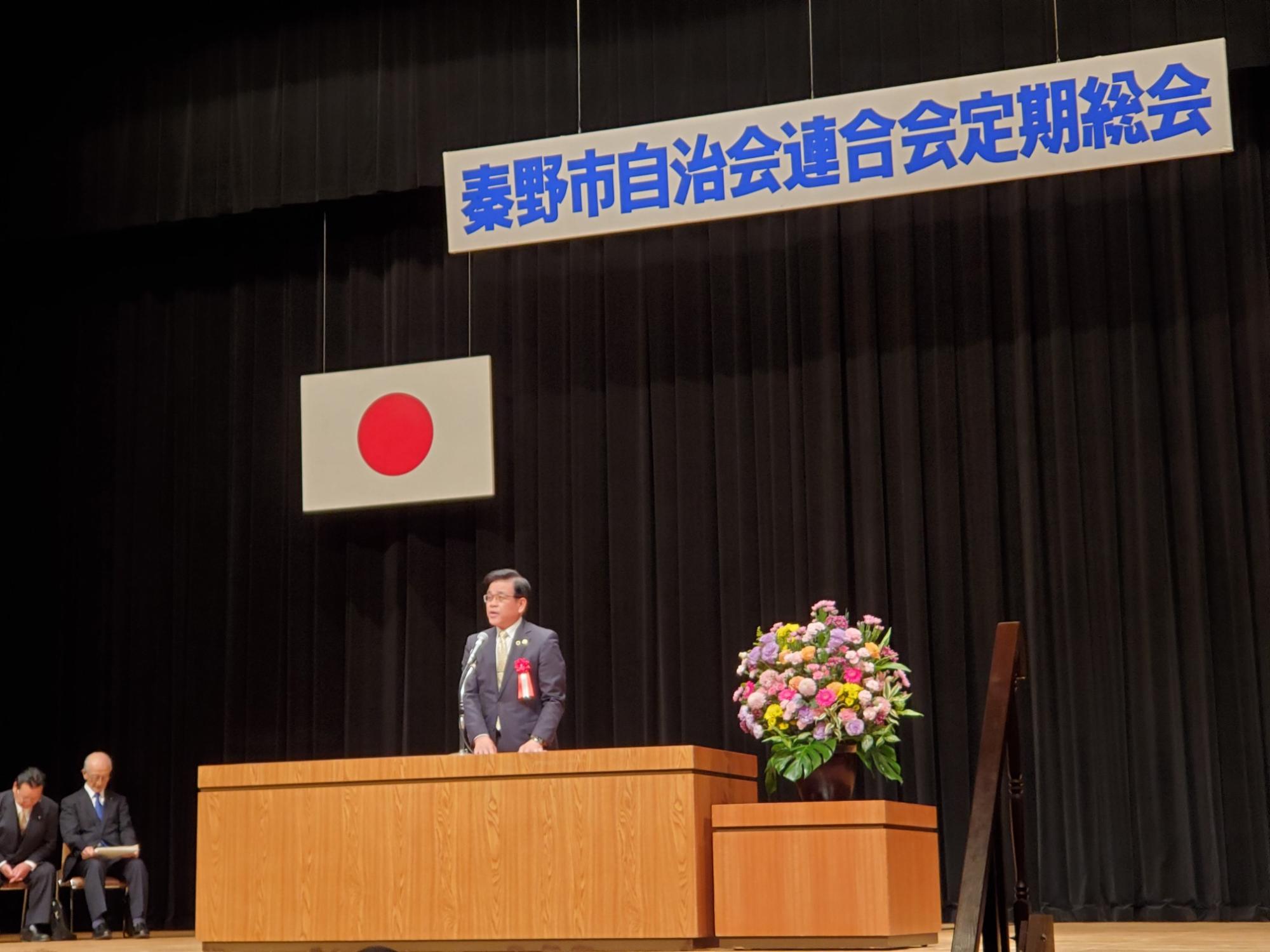 秦野市自治会連合会定期総会の看板と日の丸が掲げられたステージ上で、スーツ姿の市長が演台に立ち、マイクでスピーチをしており、その横には豪華な花束が飾られている写真