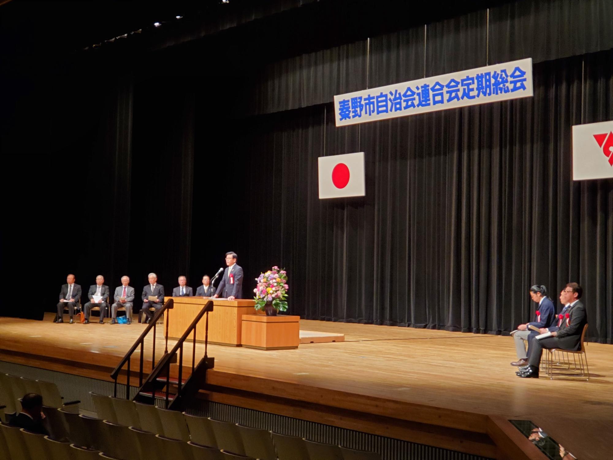 秦野市自治会連合会定期総会の看板と日の丸が掲げられたステージ上で、スーツ姿の市長が演台に立ち、マイクでスピーチをしており、壇上には数人の関係者が着席している様子の写真