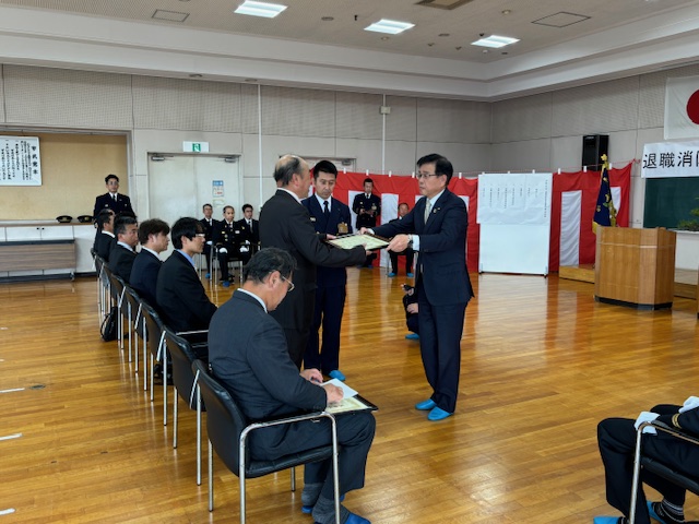 広間で行われている退職消防団員感謝状贈呈式で、スーツ姿の市長が男性に感謝状を手渡している様子の写真