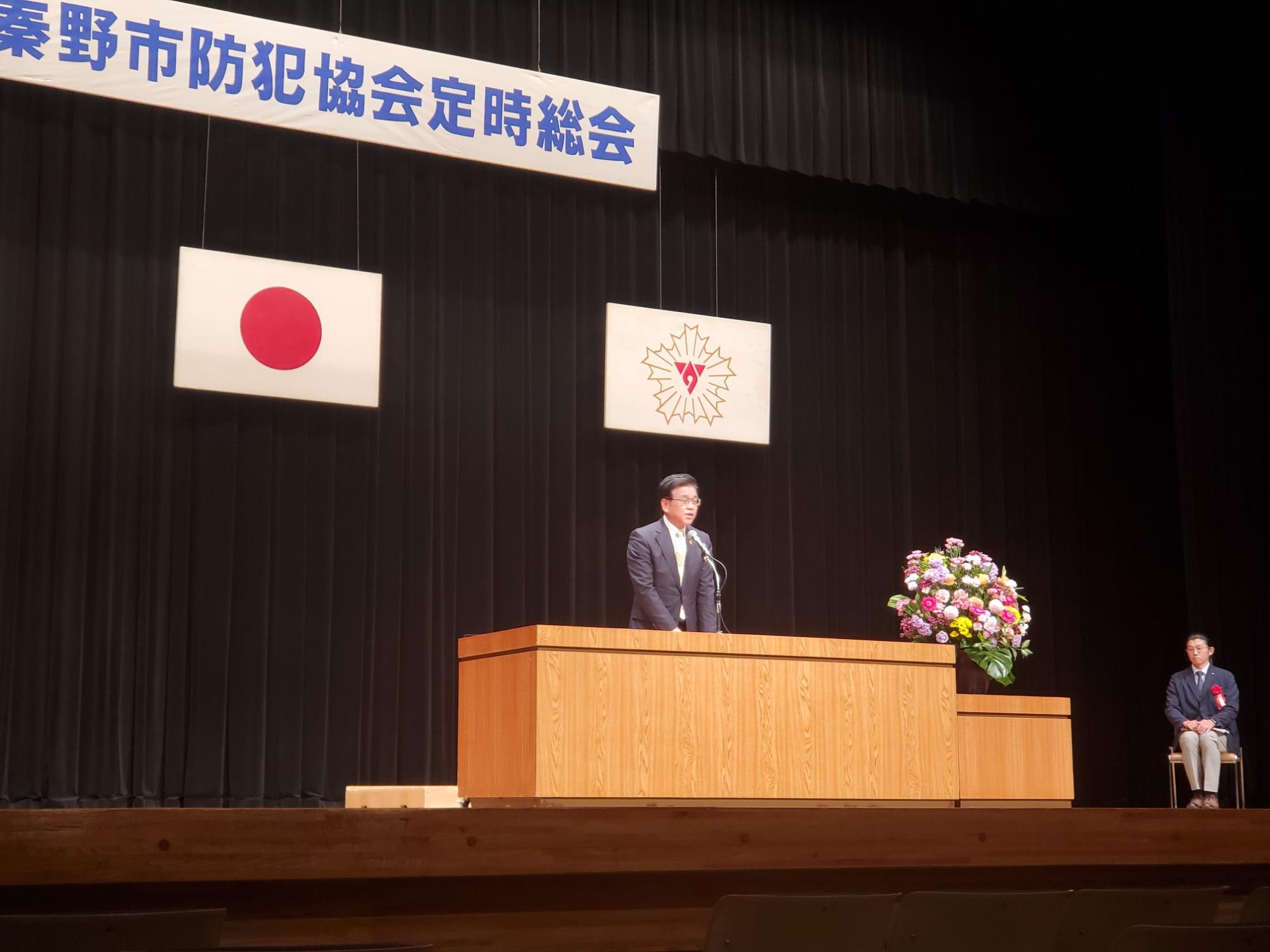 秦野市防犯協会定時総会と書かれた横断幕と日の丸の下、ステージ上の演台に立つスーツ姿の市長がスピーチをしており、右側には花束と椅子に座る男性が写っている写真