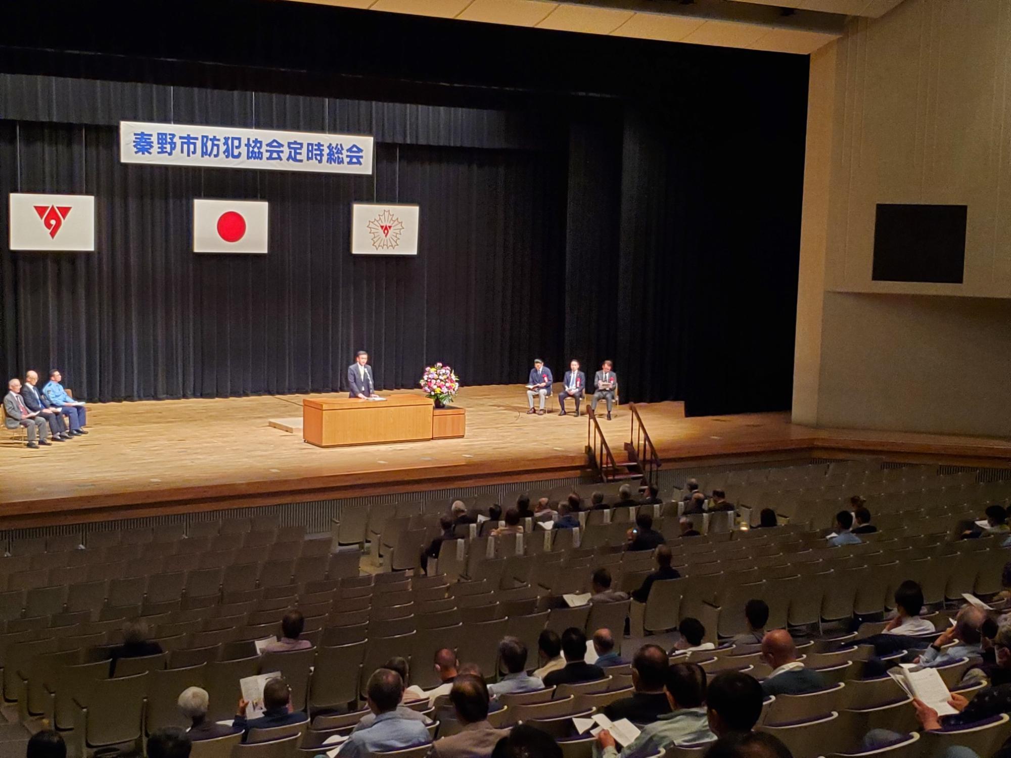 秦野市防犯協会定時総会の横断幕と日の丸の下、ステージ上の演台に立つスーツ姿の市長がスピーチをしており、複数の人が壇上に、大勢の聴衆が客席に座っている様子の写真