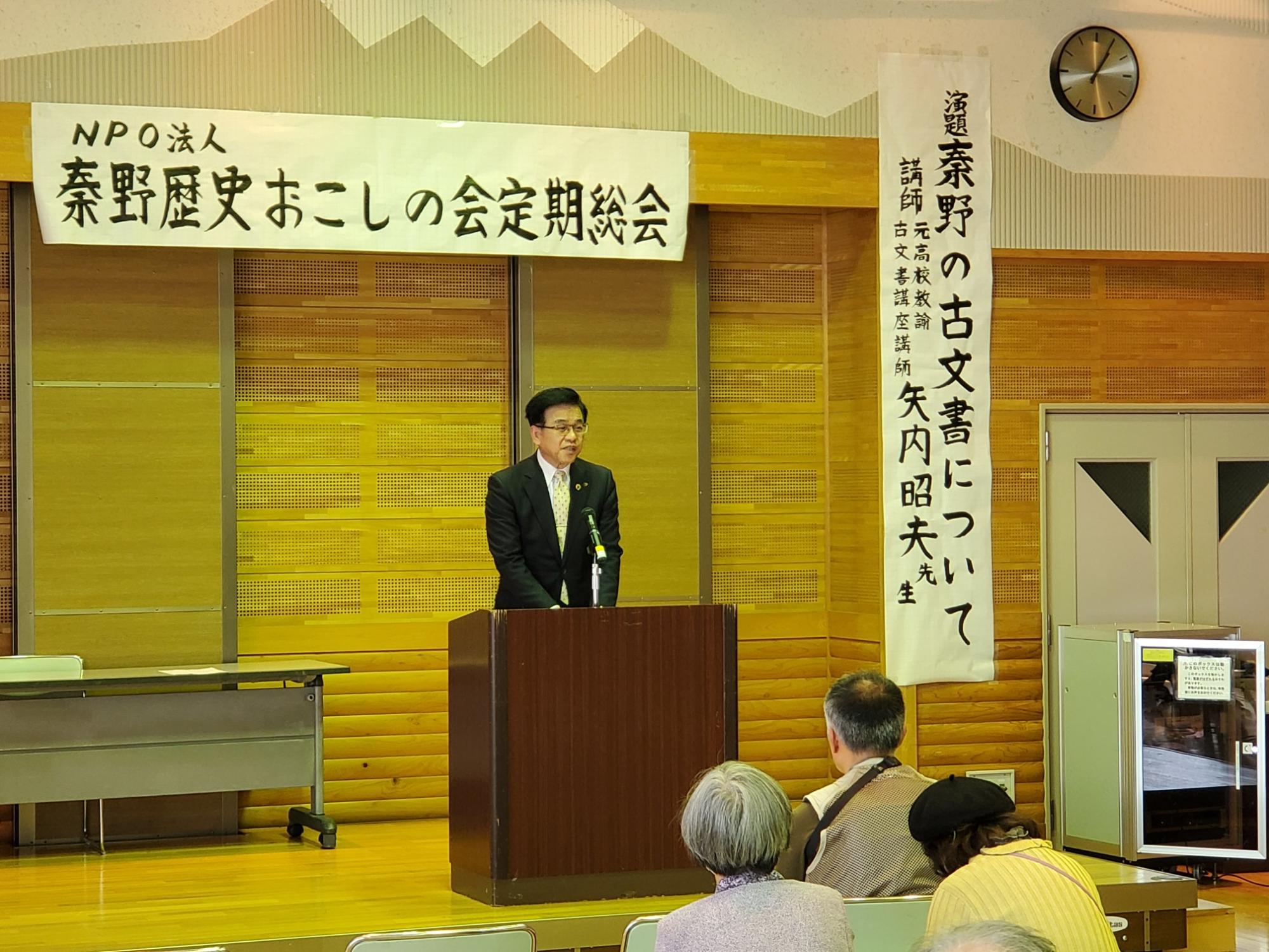 「NPO法人 秦野歴史おこしの会 定例総会」と書かれた横断幕がある会場で、スーツを着た市長が演台に立ち、大勢の聴衆に向かってスピーチをしている様子の写真