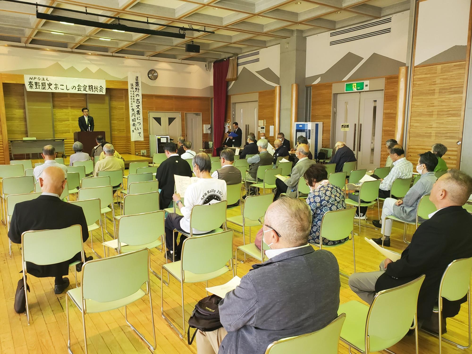 「NPO法人 秦野歴史おこしの会 定例総会」と書かれた横断幕の下で、スーツを着た市長が演台から大勢の聴衆に向かってスピーチをしている様子の写真