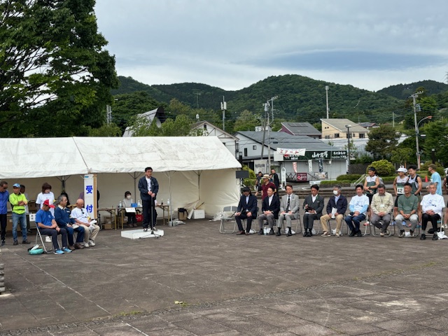 屋外でスーツを着た市長が演台に立ち、白いテントや山を背景に、椅子に座った人々や立っている人々に向かってスピーチをしている様子の写真